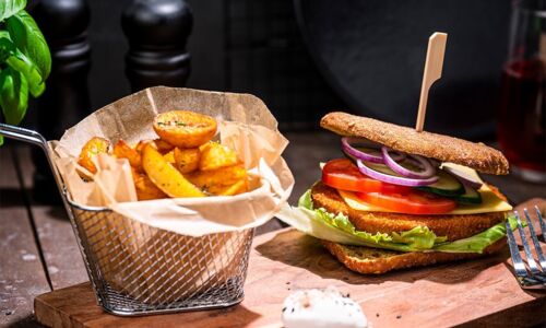 Vegane Crispy-Burger auf Holzbrett mit Gemüse, Salat und Pommes in Metallkorb, dekoriert mit Basilikum und Gewürzen, auf rustikalem Holztisch