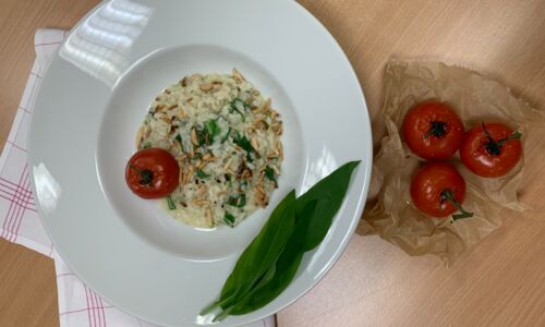 Veganes Bärlauchrisotto mit gerösteten Pinienkernen und Tomate, angerichtet auf weißem Teller, daneben frische Tomaten auf Backpapier.