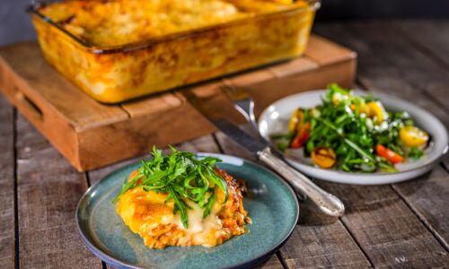 Vegane Lasagne mit veganem Hack, cremiger Sauce und geschmolzenem veganem Käseersatz, serviert mit frischem Rucola und buntem Salat auf rustikalem Holztisch.