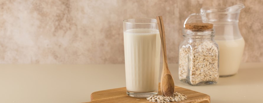 Ein Glas veganer Haferdrink auf einem Holzbrett, daneben ein Löffel mit Haferflocken und ein Glasbehälter im Hintergrund. Ideal für gesundes veganes Leben!