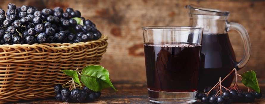 Ein Glas veganer Holundersaft neben einem Krug und einem Korb voller Holunderbeeren auf Holztisch, ideal für natürliche Erfrischung.