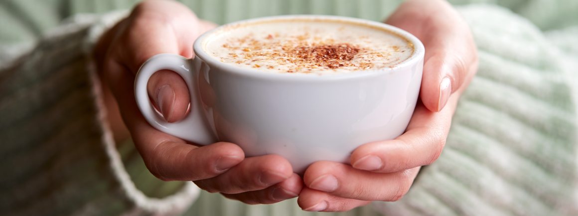 Weiße Tasse mit cremigem veganem Cappuccino in den Händen gehalten, mit einer Prise Kakaopulver bestäubt. Genuss pur!