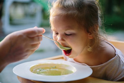 Ein Kind isst aus einem weißen Teller eine grüne vegane Suppe, im Hintergrund ein unscharfer Garten.