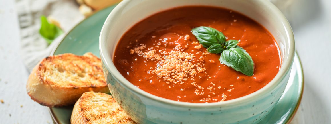 Eine Schüssel mit veganer Tomatensuppe, garniert mit Basilikum und Brotkrusten auf einem Teller mit geröstetem Brot.