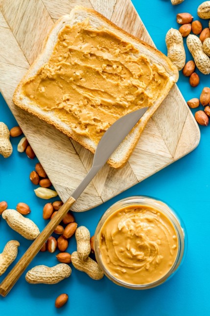 Ein Glas vegane Erdnussbutter neben einem mit Erdnussbutter bestrichenen Toast und Erdnüssen auf einem Schneidebrett.