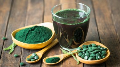 Ein Glas grüner Spirulina-Smoothie, Spirulina-Pulver und Spirulina-Tabletten auf Holzlöffeln, auf einem Holztisch.