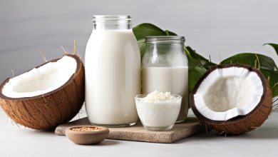 Geöffnete Kokosnüsse, zwei Gläser mit Kokosmilch und eine Schüssel Kokosraspeln auf einem Holzbrett vor einem grünen Blatt.